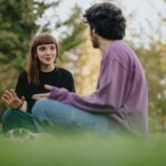 Two young adults sitting on the grass in a park, engaged in a thoughtful conversation, surrounded by trees and soft afternoon light.