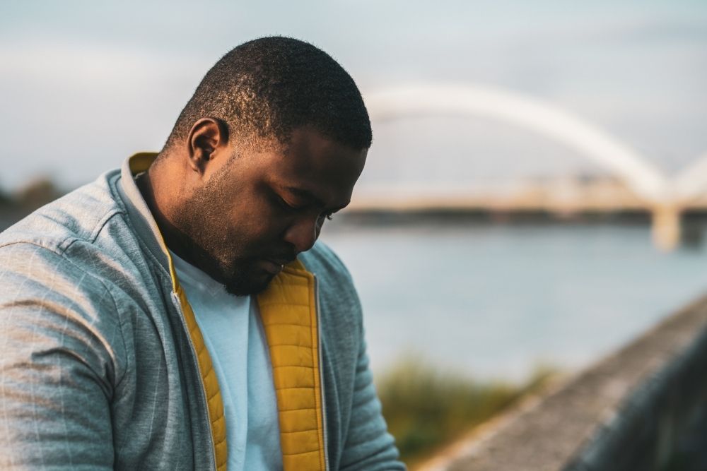 Man standing outdoors near a body of water, looking down with a serious expression