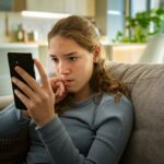 A concerned teenage girl sits on a couch, looking anxiously at her phone, with her hand near her mouth, suggesting worry or distress.