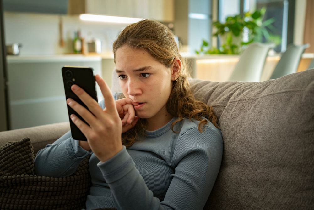 A concerned teenage girl sits on a couch, looking anxiously at her phone, with her hand near her mouth, suggesting worry or distress.