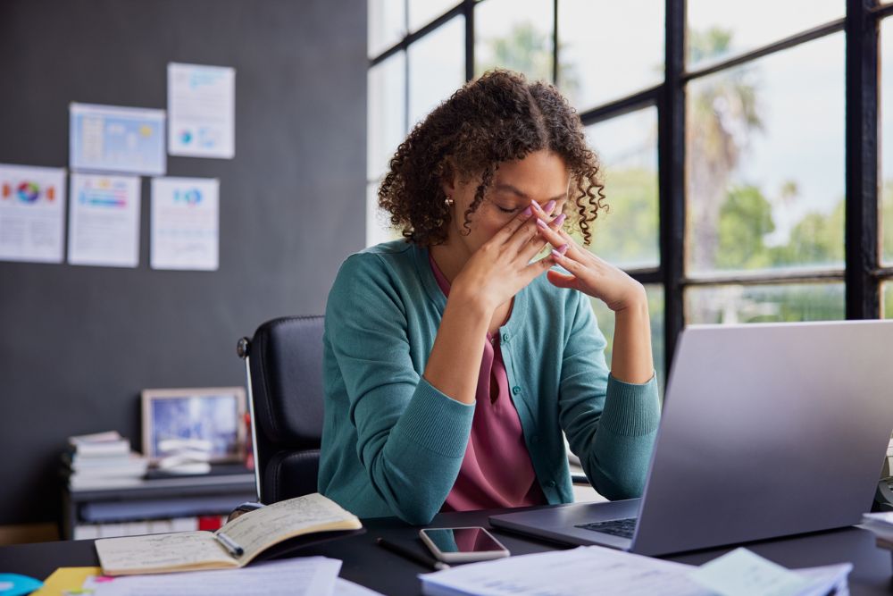 Woman sitting at a desk with her eyes closed and fingers pressed to her forehead, showing signs of stress or burnout in an office setting.