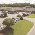 An aerial view of Carolina Dunes Behavioral Health, a large brick building with multiple wings surrounded by a parking lot full of cars. Trees and landscaped greenery border the property.