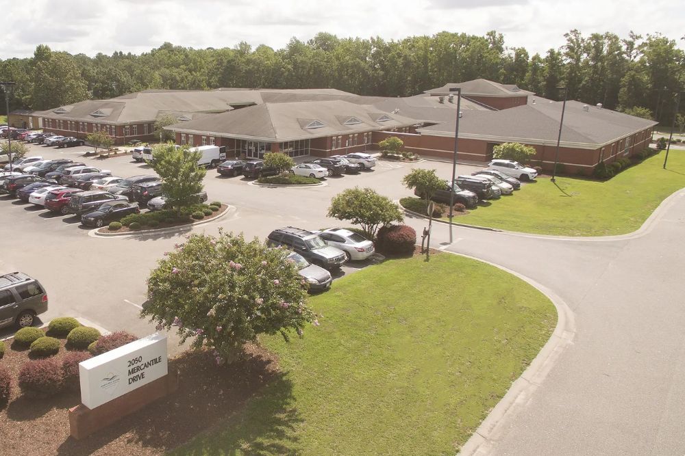 An aerial view of Carolina Dunes Behavioral Health, a large brick building with multiple wings surrounded by a parking lot full of cars. Trees and landscaped greenery border the property.