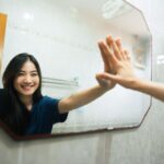 Smiling woman standing in front of a bathroom mirror, touching her reflection with a high-five gesture, symbolizing self-affirmation and confidence.