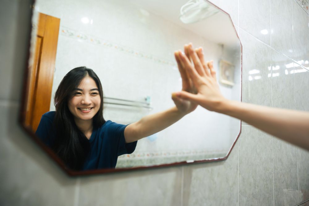 Smiling woman standing in front of a bathroom mirror, touching her reflection with a high-five gesture, symbolizing self-affirmation and confidence.