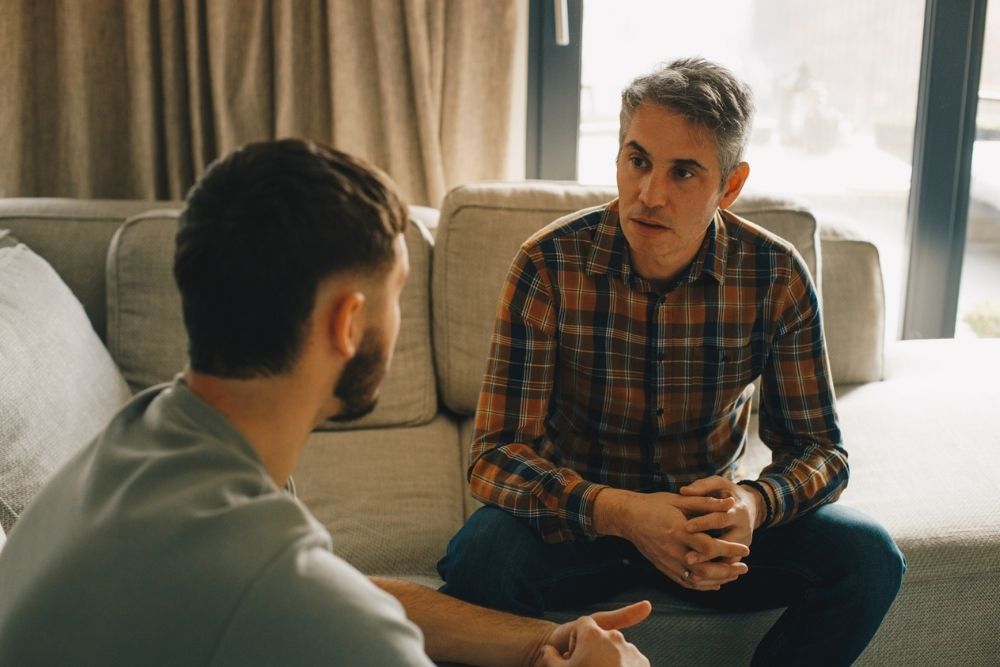 Two men sitting on a couch having a serious conversation, suggesting an open discussion about boundaries or personal challenges.