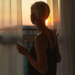 Woman holding a glass while looking out a window at the sunset through curtains.