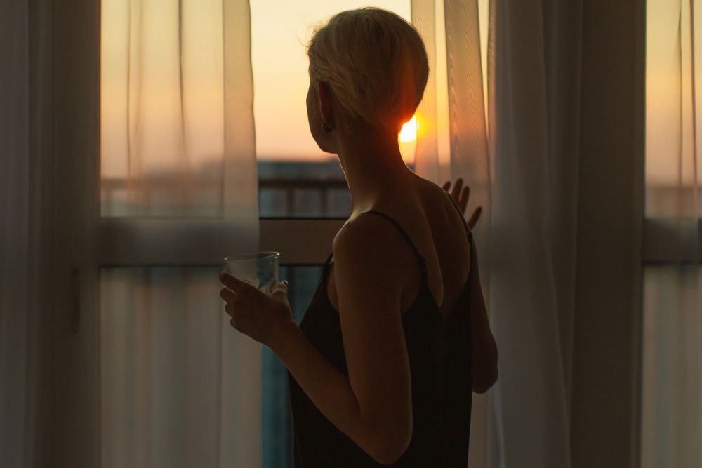 Woman holding a glass while looking out a window at the sunset through curtains.