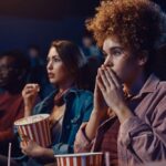 Group of people sitting in a movie theater watching a scary film, looking tense and focused while holding popcorn and drinks.
