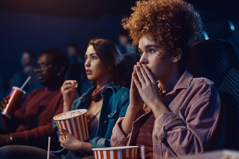 Group of people sitting in a movie theater watching a scary film, looking tense and focused while holding popcorn and drinks.