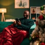 A man relaxes on a couch with a red blanket, smiling as he looks at his phone. A decorated Christmas tree glows beside him in a cozy, festive living room.