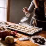 A woman dusts powdered sugar over a tray of cookies in a warm, cozy kitchen, surrounded by holiday ingredients and baking supplies.