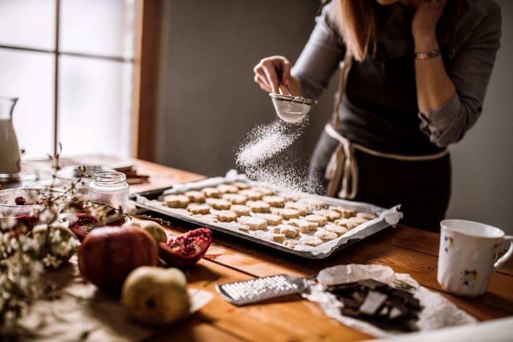 A woman dusts powdered sugar over a tray of cookies in a warm, cozy kitchen, surrounded by holiday ingredients and baking supplies.