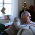An older man sits on a couch, looking out a window with a thoughtful expression in a softly decorated home during the holidays.
