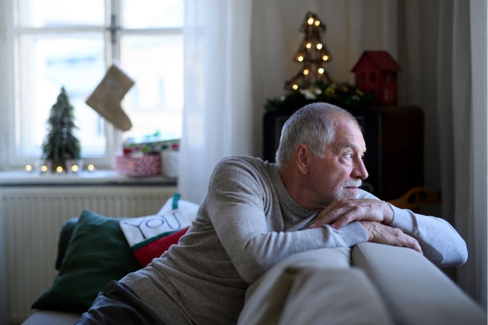 An older man sits on a couch, looking out a window with a thoughtful expression in a softly decorated home during the holidays.