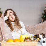 A tired or stressed teenage girl sitting on a couch during the holidays, resting her head on her hand, with a decorated Christmas tree in the background and a small dog beside her.
