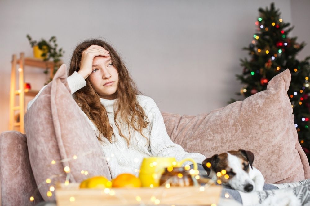 A tired or stressed teenage girl sitting on a couch during the holidays, resting her head on her hand, with a decorated Christmas tree in the background and a small dog beside her.