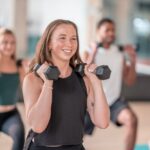 Carolina Dunes - How Exercise Promotes Mental Health. A woman in a black shirt lifts weights during a class as another woman and man do the same in the foreground.