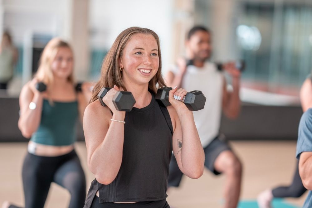 Carolina Dunes - How Exercise Promotes Mental Health. A woman in a black shirt lifts weights during a class as another woman and man do the same in the foreground.