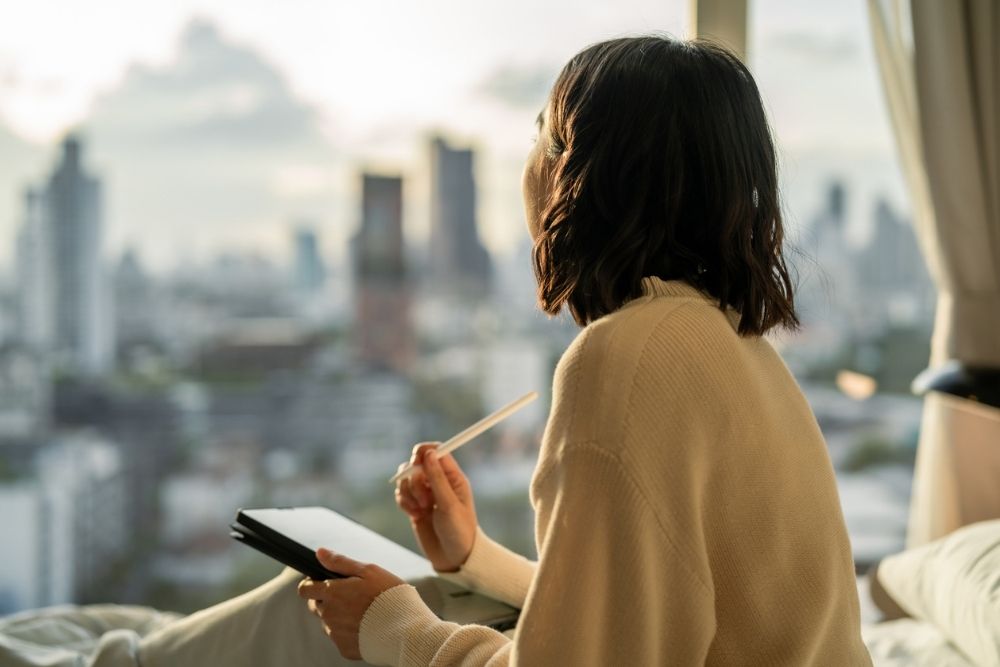 Person sitting by a window, journaling and looking out at a city skyline, reflecting quietly in a calm indoor setting.